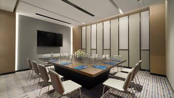 A conference room with a large rectangular table, white chairs, a flat-screen TV, and a patterned carpet.