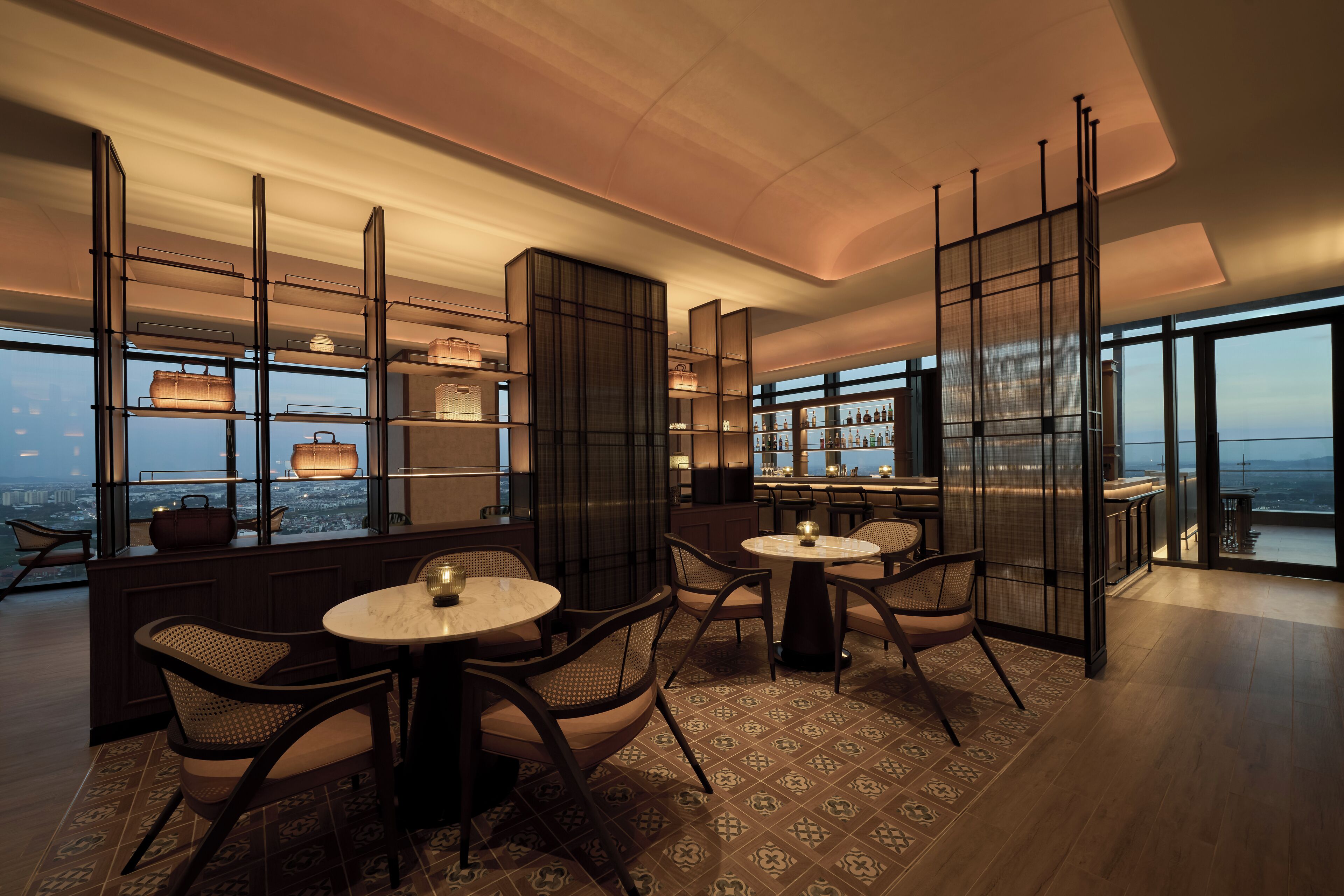 A modern hotel lobby with high ceilings, large windows, and a view of the cityscape. There are round tables and chairs arranged for guests.