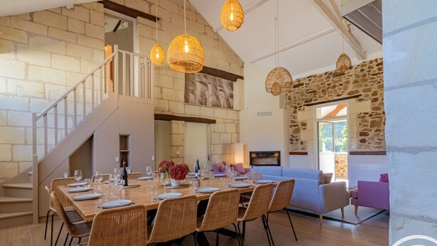 A spacious dining area with a long wooden table set for six, surrounded by wicker chairs. A staircase with a white railing leads to an upper level. - Spacious 8-bedroom cottage in Anjou vineyards (Blaison-Saint-Sulpice)