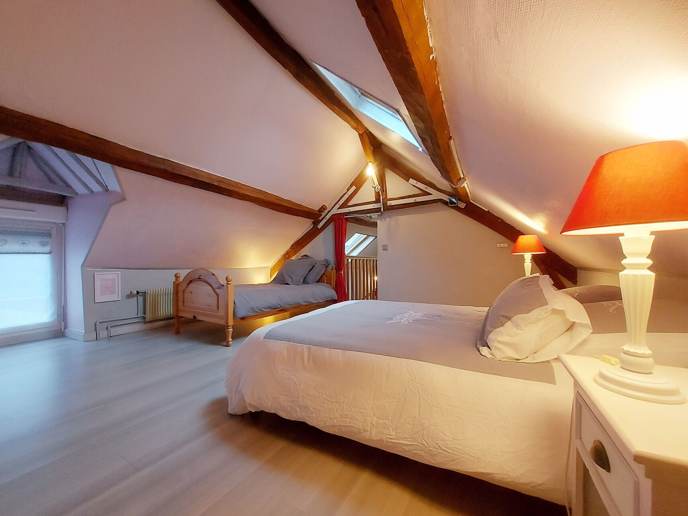 Een slaapkamer met een schuine kap, een bed met witte beddengoed, een nachtkastje met een lamp en een raam met uitzicht op het omliggende landschap.