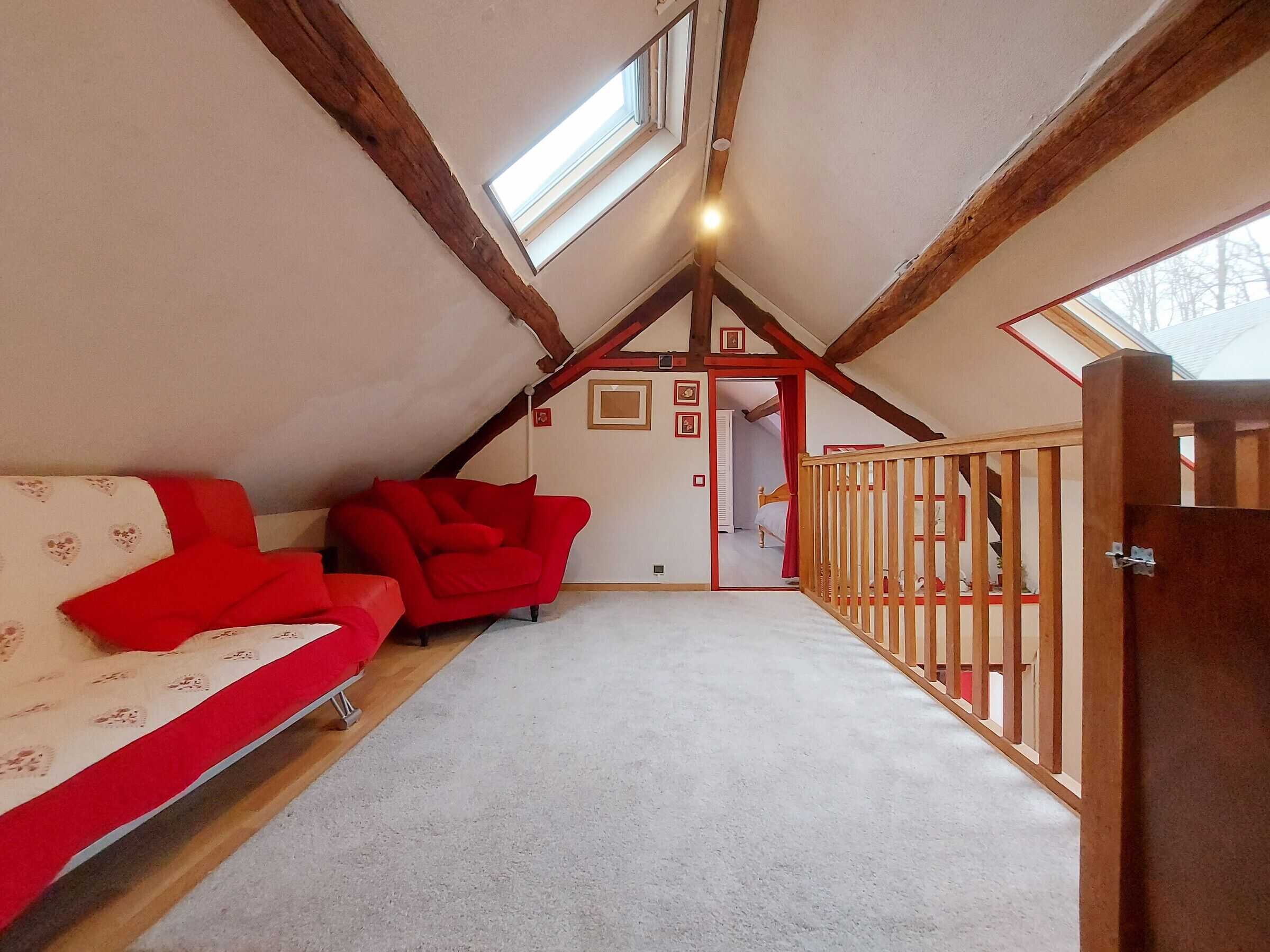 A cozy attic bedroom with a sloped ceiling, wooden beams, a red sofa, a white patterned couch, and a view through a door to another room.