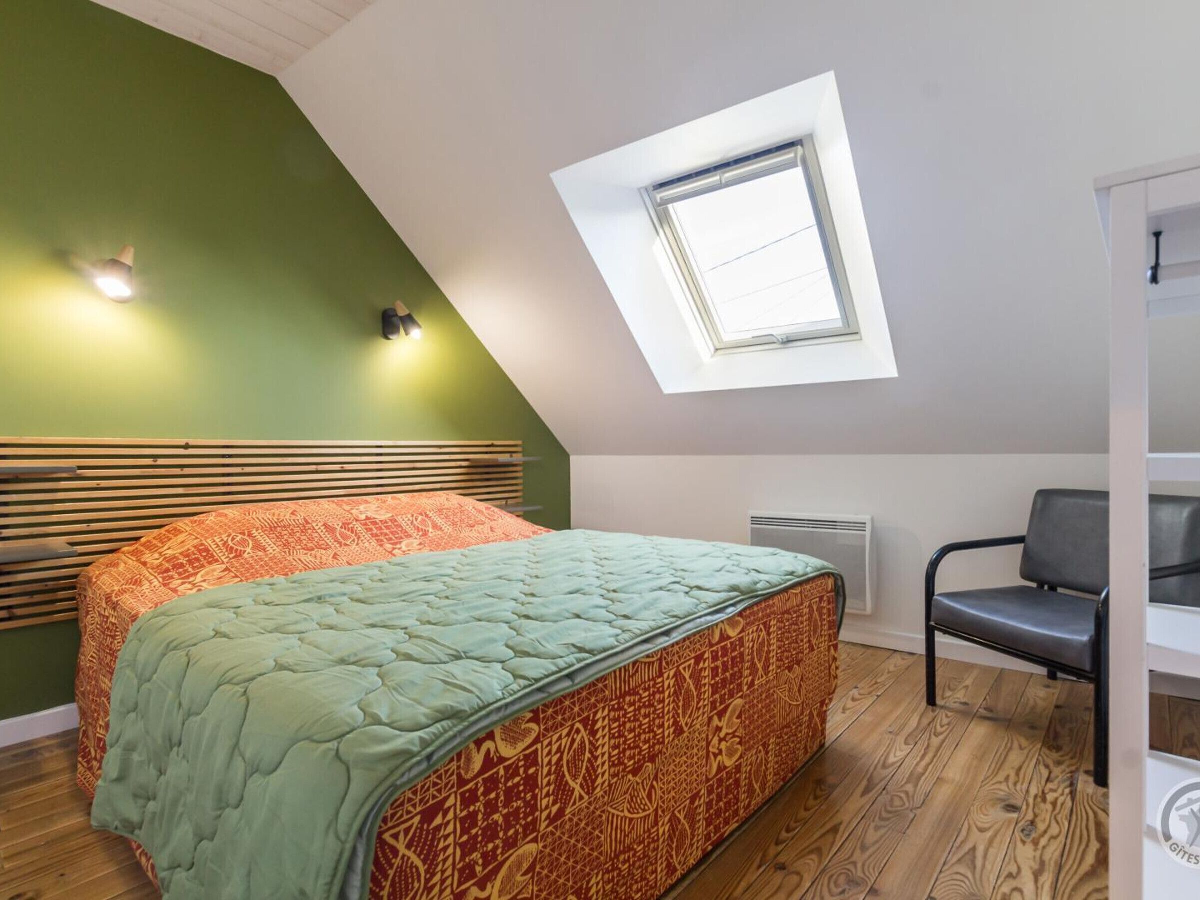 A bedroom with a sloped ceiling, a skylight, a bed with patterned bedding, a chair, and a radiator.