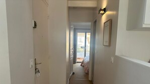 Un pasillo con piso alfombrado, una puerta a la izquierda y una cama a lo lejos.