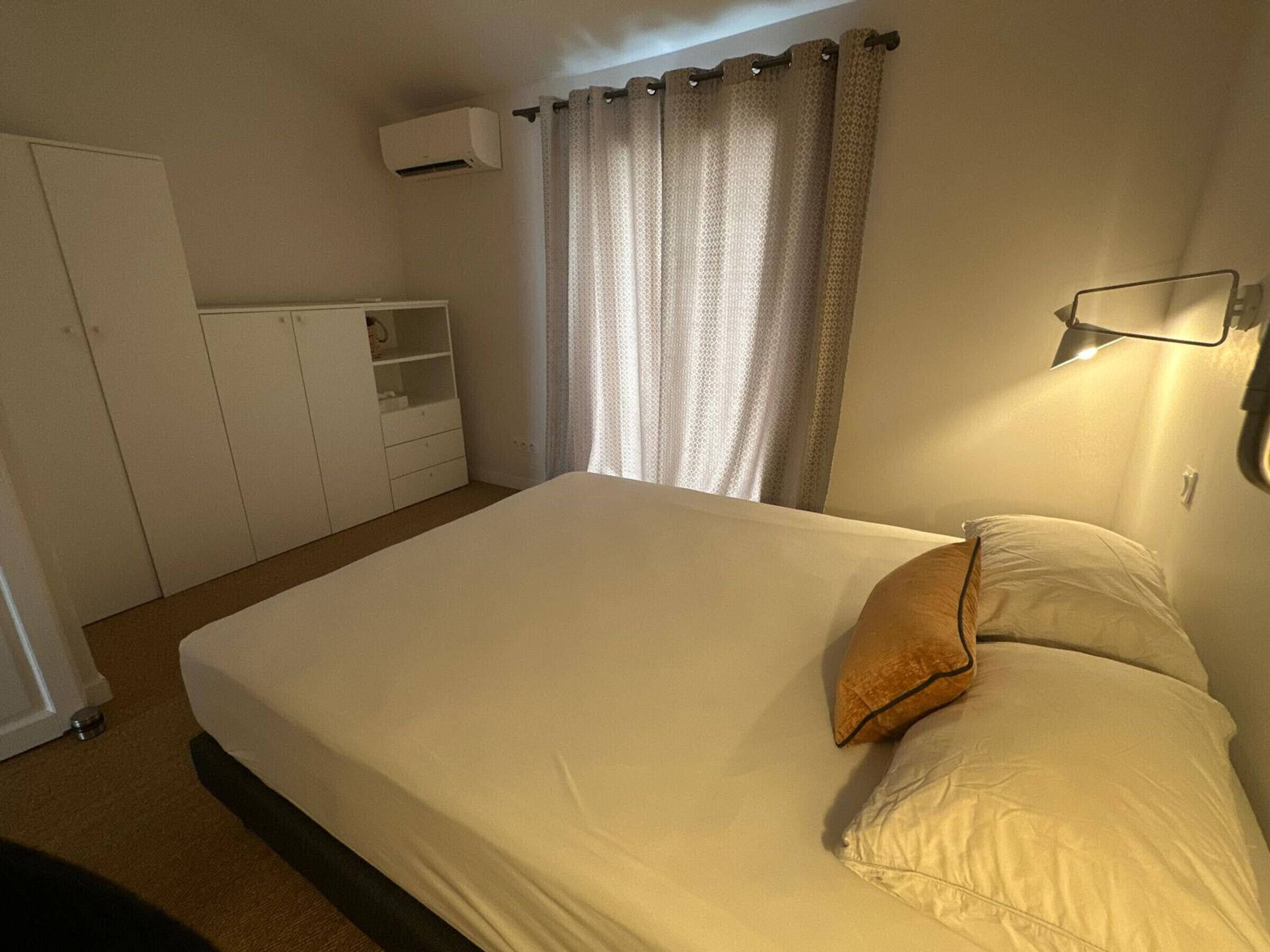 A bedroom with a bed, a wardrobe, a bedside table, a lamp, and a window with curtains.