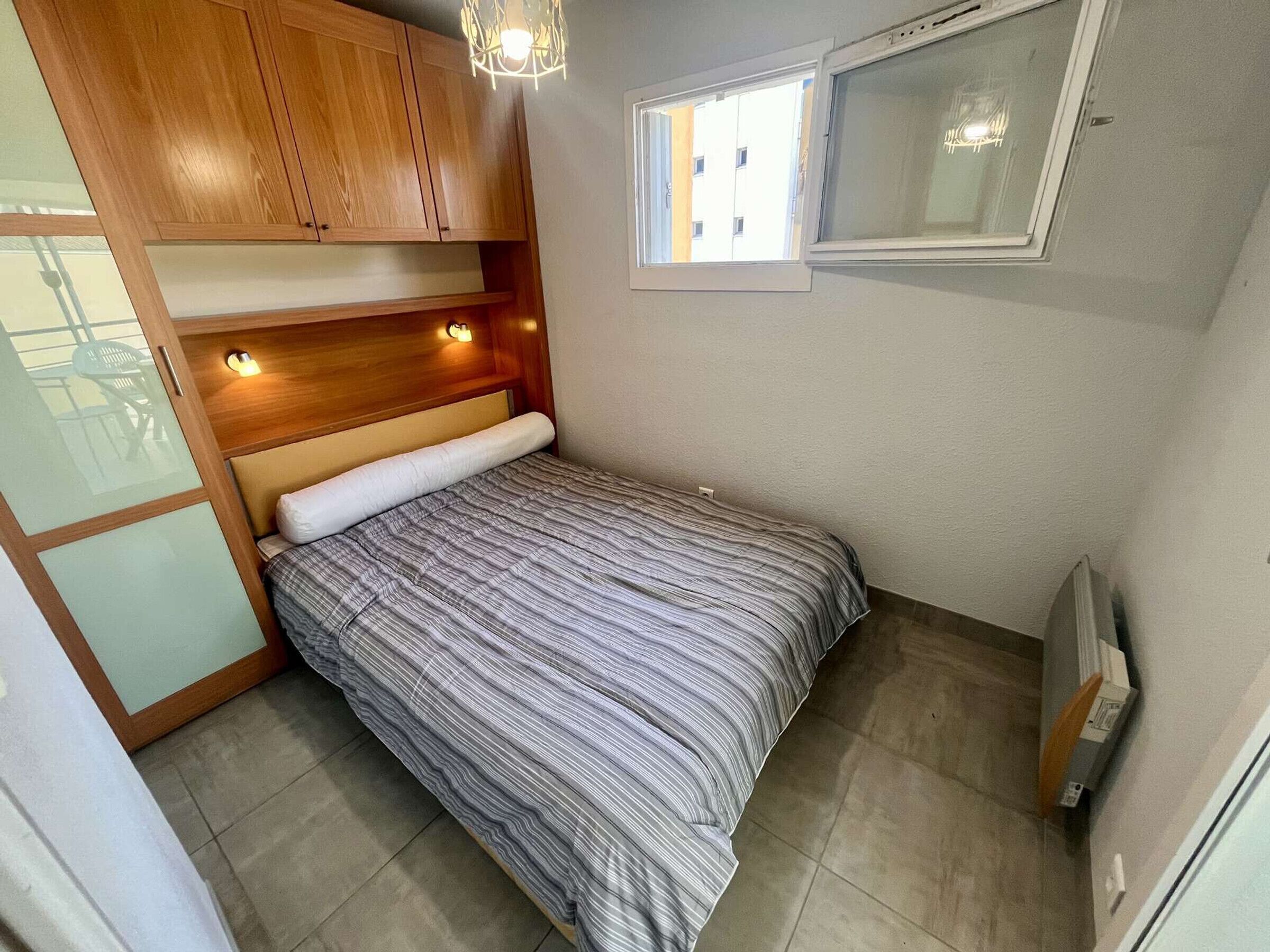 A compact bedroom with a bed, wooden cabinets, a window, and a ceiling light fixture.