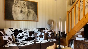 Een woonkamer met een bank met koeienhuidprint, een schilderij van een wolf in een lijst, een houten trap en een rond houten bijzettafeltje met kaarsen.