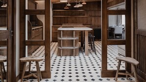 Un intérieur rustique avec des poutres apparentes, une grande table en bois au centre, entourée de tabourets, et un sol à motifs.