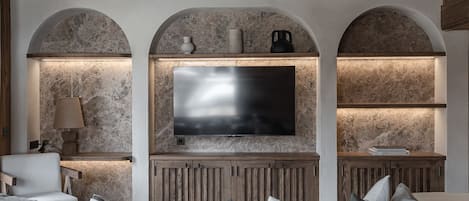 A bedroom with a large bed, a TV mounted on a stone wall, wooden shelves, and a lamp.