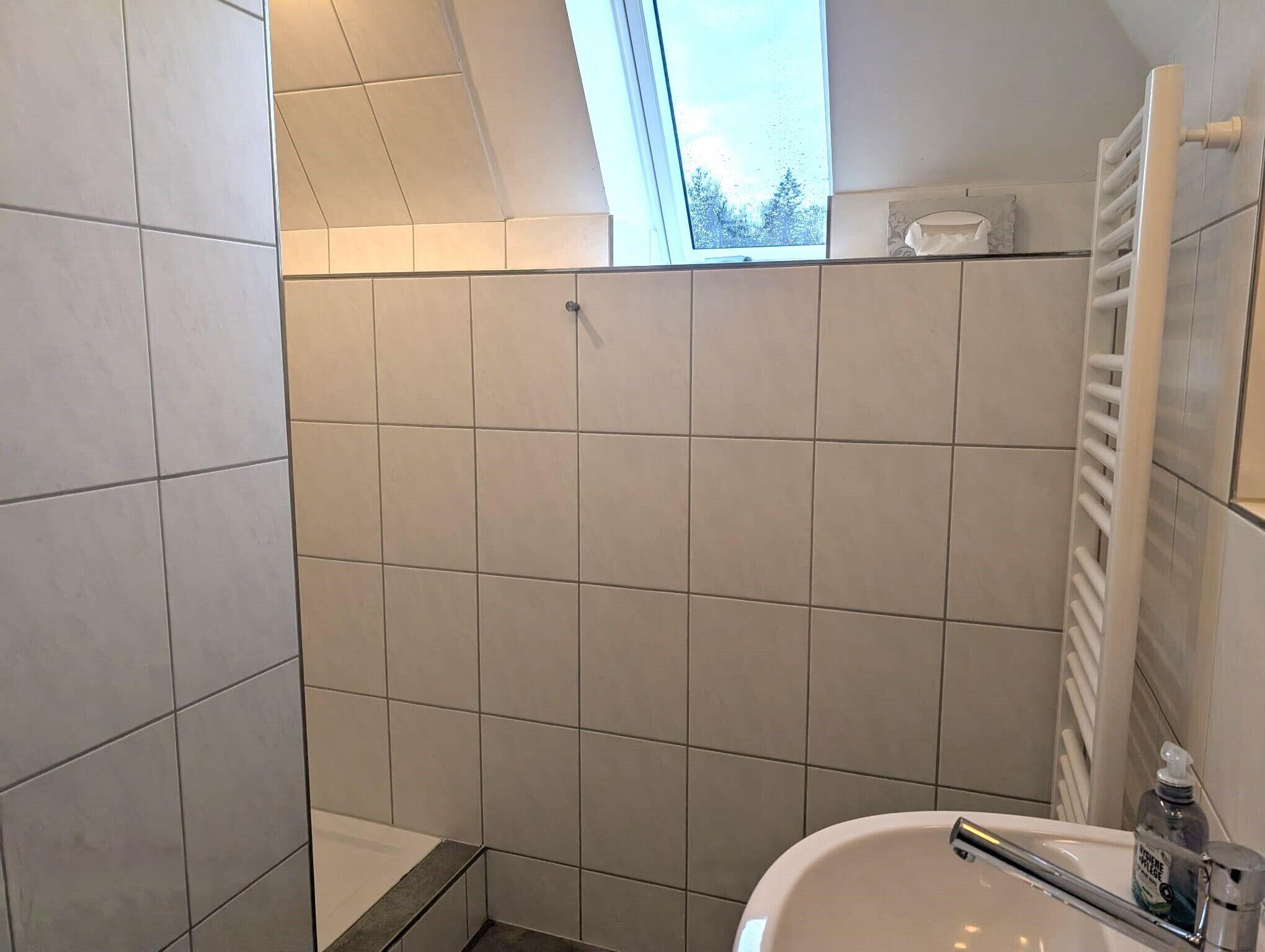 Uma casa de banho com azulejos brancos, uma janela, um porta-toalhas e uma pia com dispensador de sabonete.