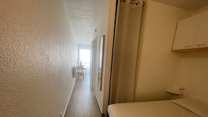 A narrow hallway with a bed on the right, a door on the left, and a table with chairs in the distance. - 2-star apartment near thermal baths with balcony (Balaruc-les-Bains)