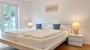 A double bed with white bedding, two nightstands with lamps, a framed picture on the wall, and a window with a view of greenery.