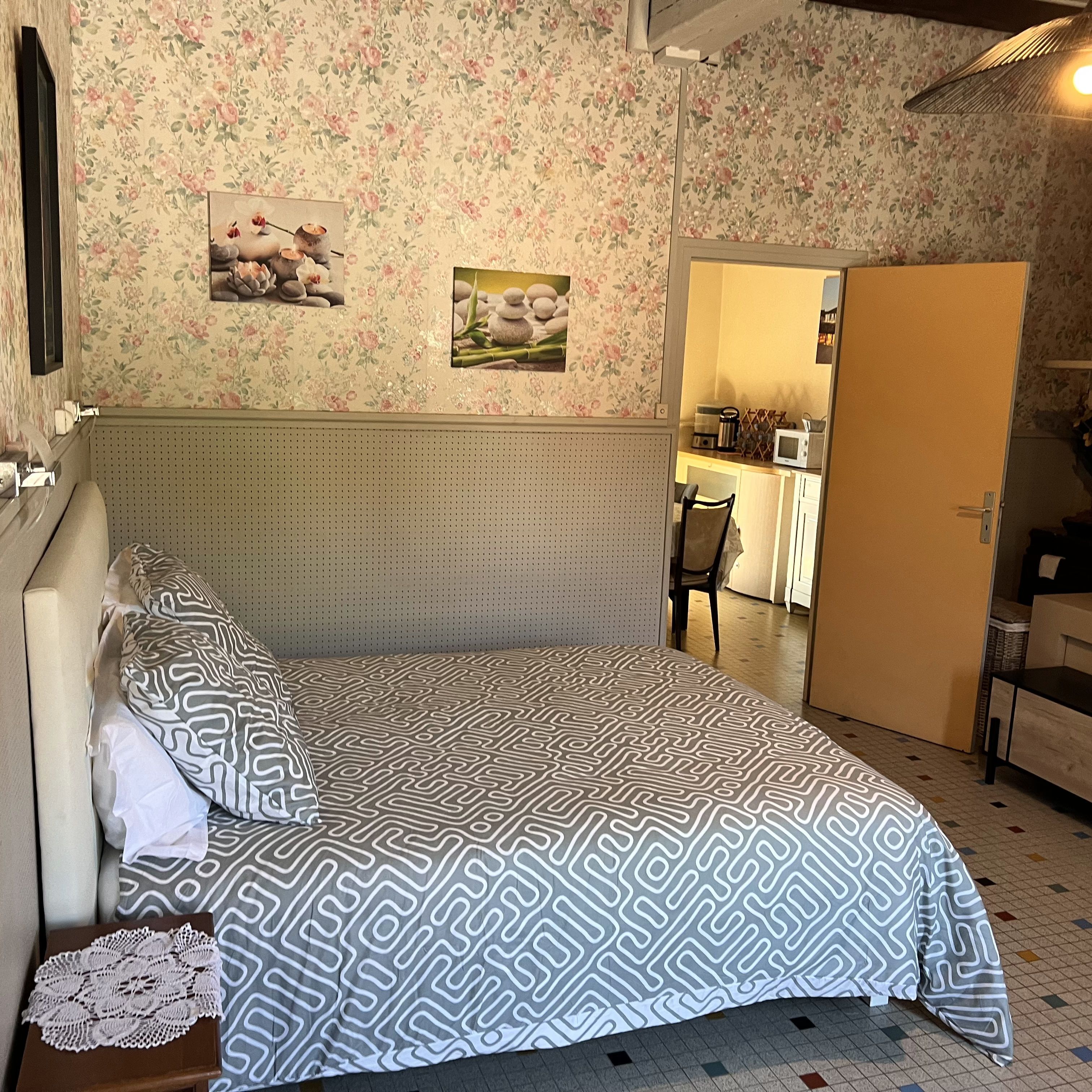 A bedroom with a bed, a wooden headboard, a floral wallpaper, and a small kitchen area visible through an open door.