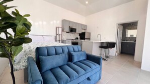 Een moderne woonkamer met een blauwe bank, een bed met een bloemenbedekking, en een keuken met grijze kastjes en een magnetron.