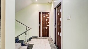 A hallway with a marble staircase, a wooden door with circular patterns, and a light switch on the wall. - Green Edge Regency (Kolkata)