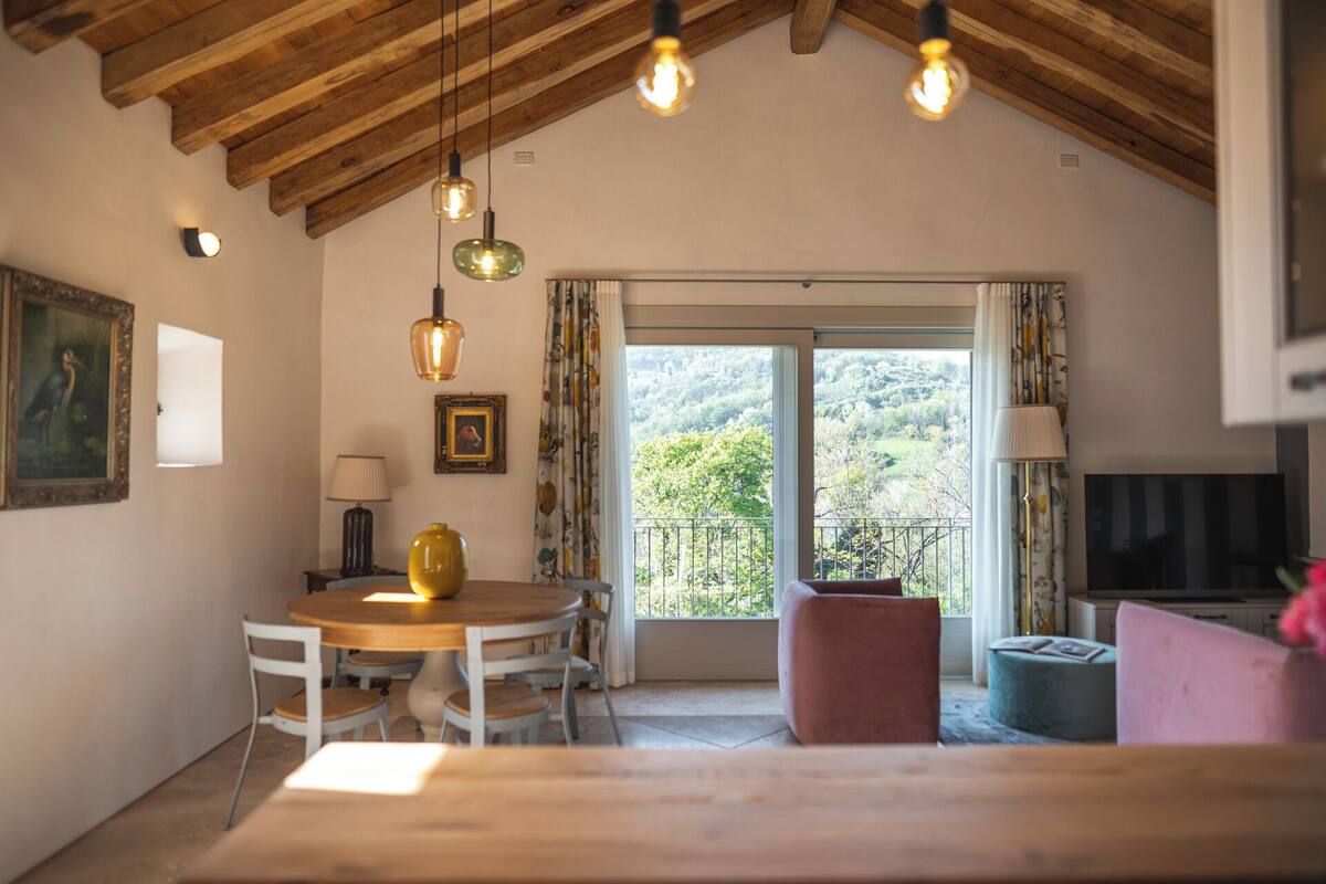 A cozy room with a wooden ceiling, a dining table with chairs, a pink sofa, and a balcony with a view of greenery.