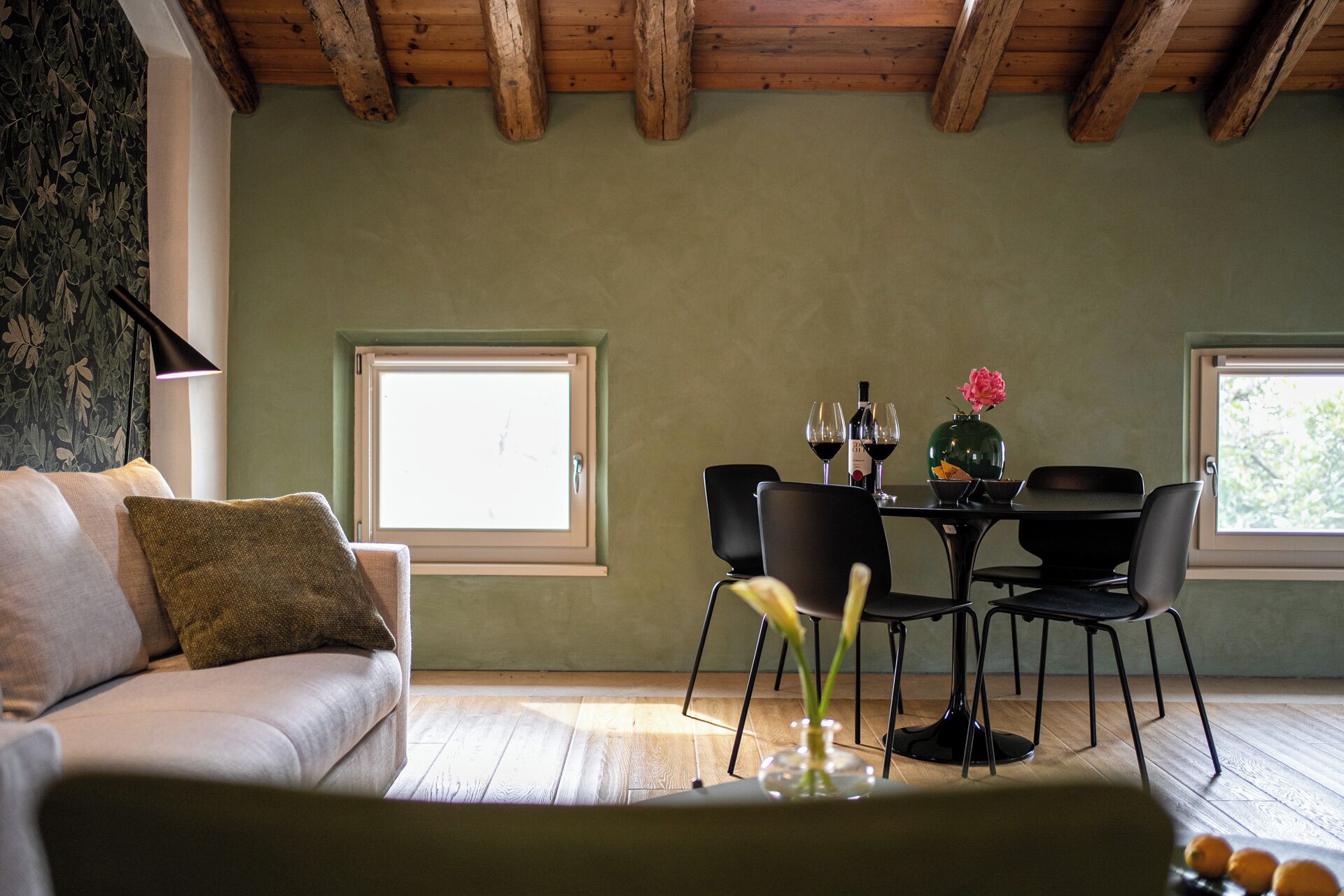 A cozy room with a wooden ceiling, a sofa with cushions, a dining table with chairs, and a window with a view.