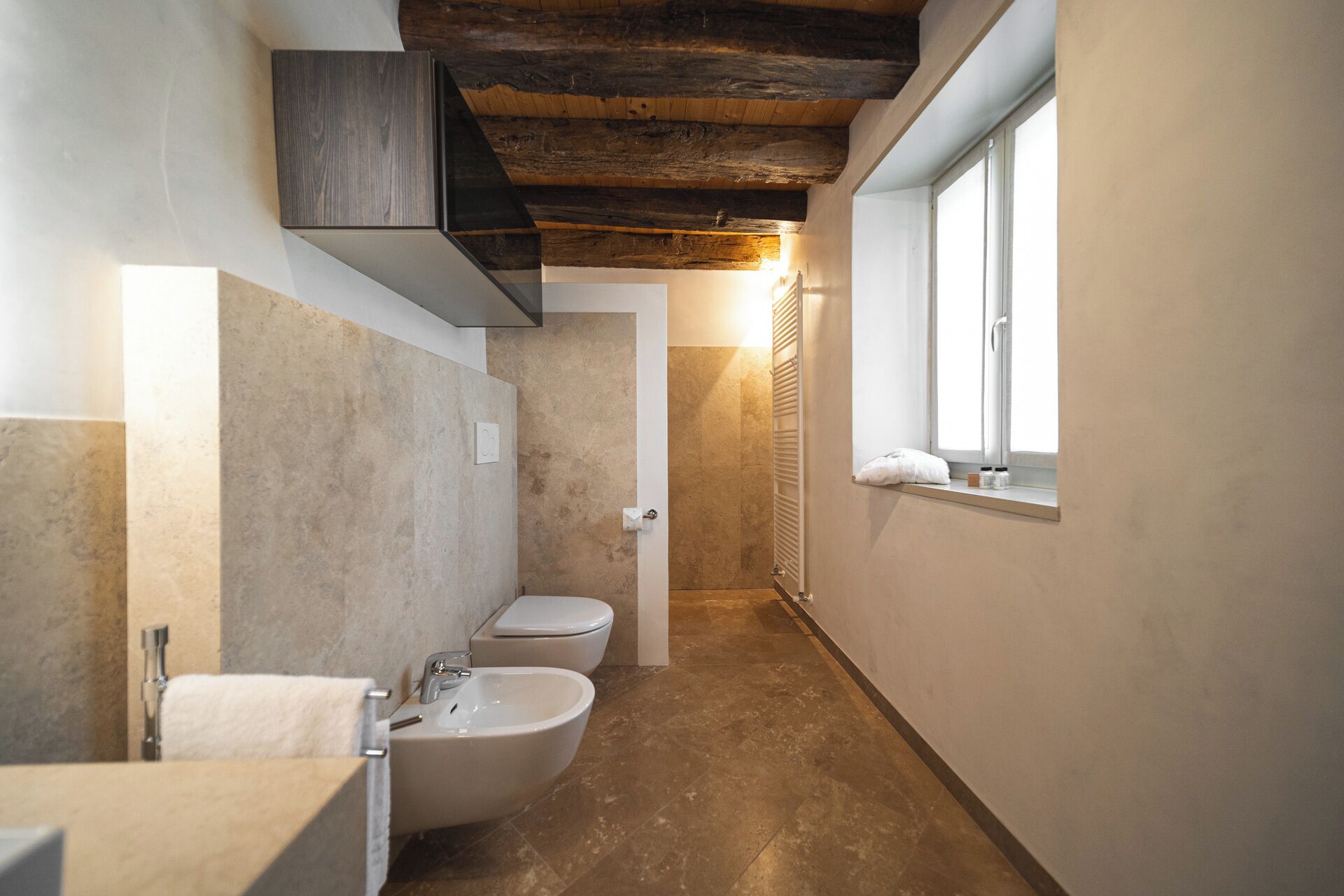 Un baño estrecho con bañera, inodoro y bidé, vigas de techo de madera y una ventana que proporciona luz natural.