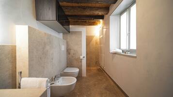 Un baño estrecho con bañera, inodoro y bidé, vigas de techo de madera y una ventana que proporciona luz natural.