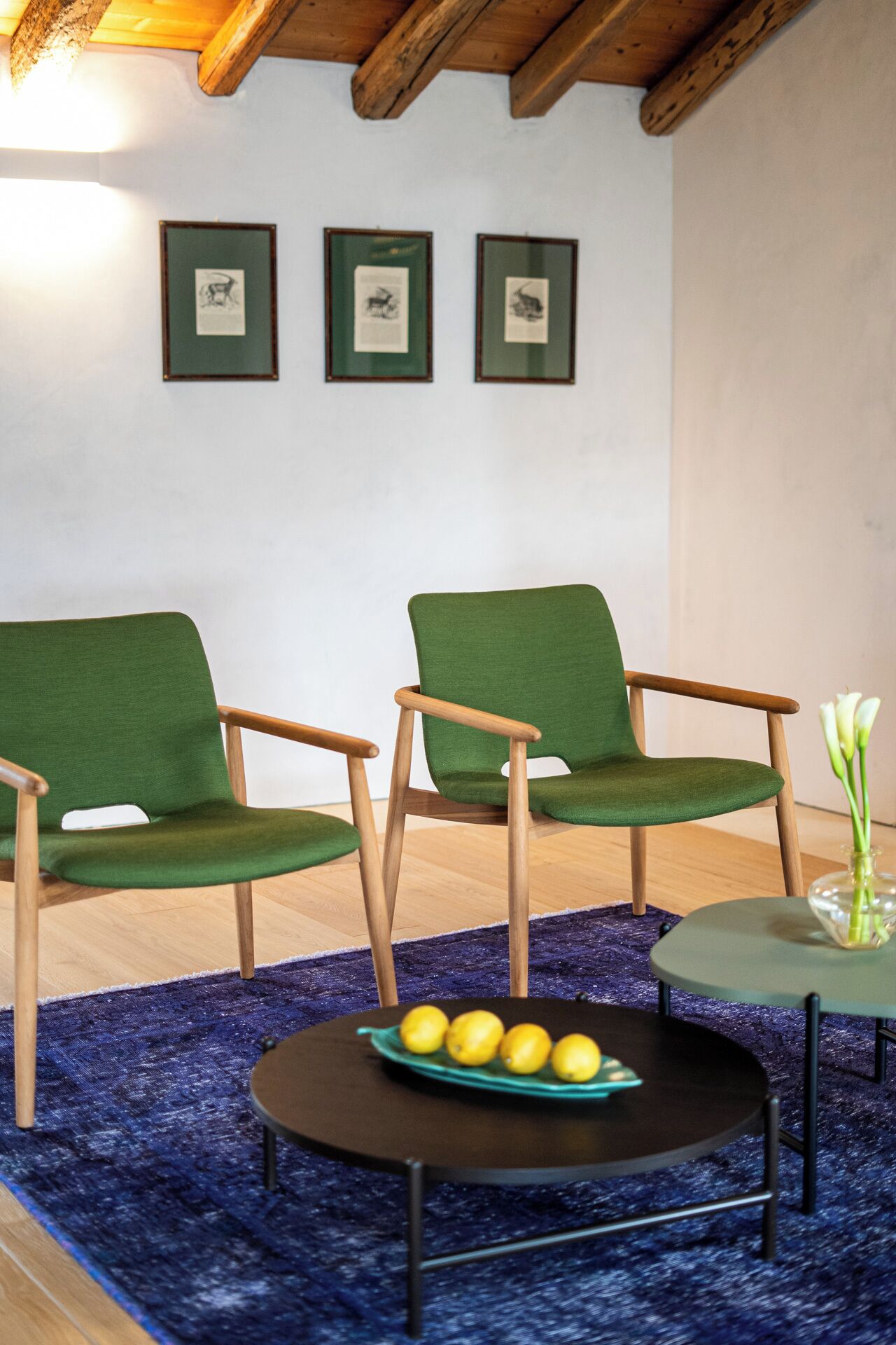 Una habitación con dos sillones tapizados en verde, una mesita redonda con limones y un jarrón con flores sobre una pequeña mesa.