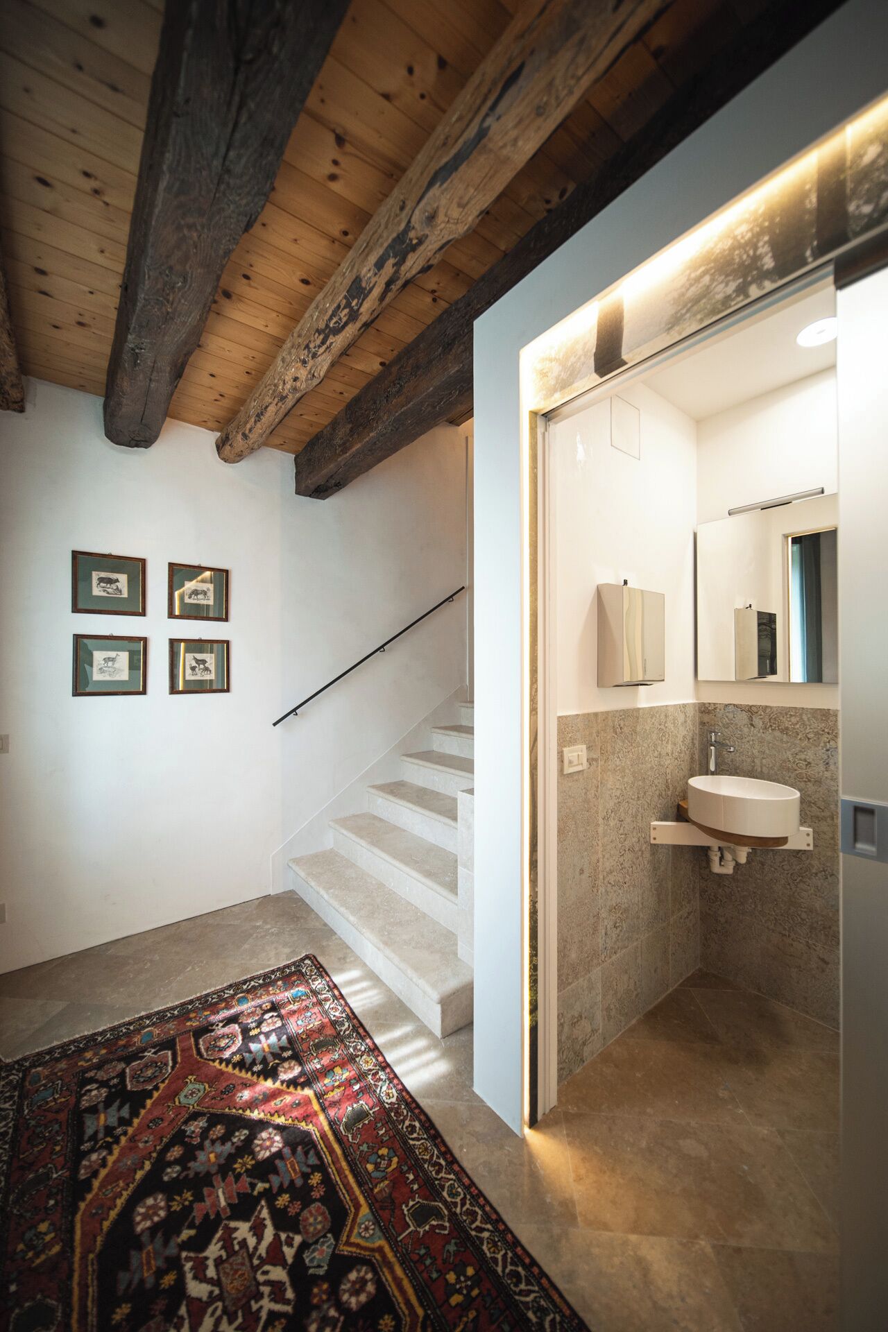 A staircase leading to a bathroom with a sink and mirror, wooden beams on the ceiling, and a patterned rug on the floor.