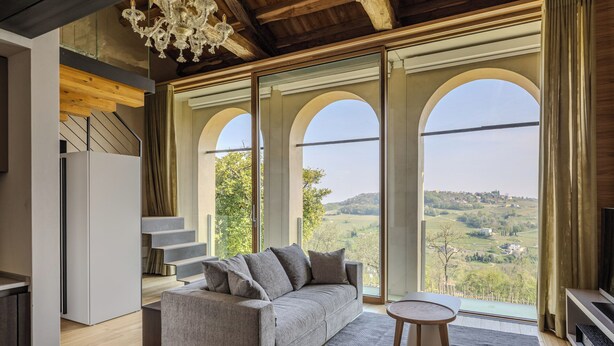 A modern living room with a sofa, a small table, and a view of a hilly landscape through large arched windows. - Tenuta Duchi Corbanus (Tarzo)