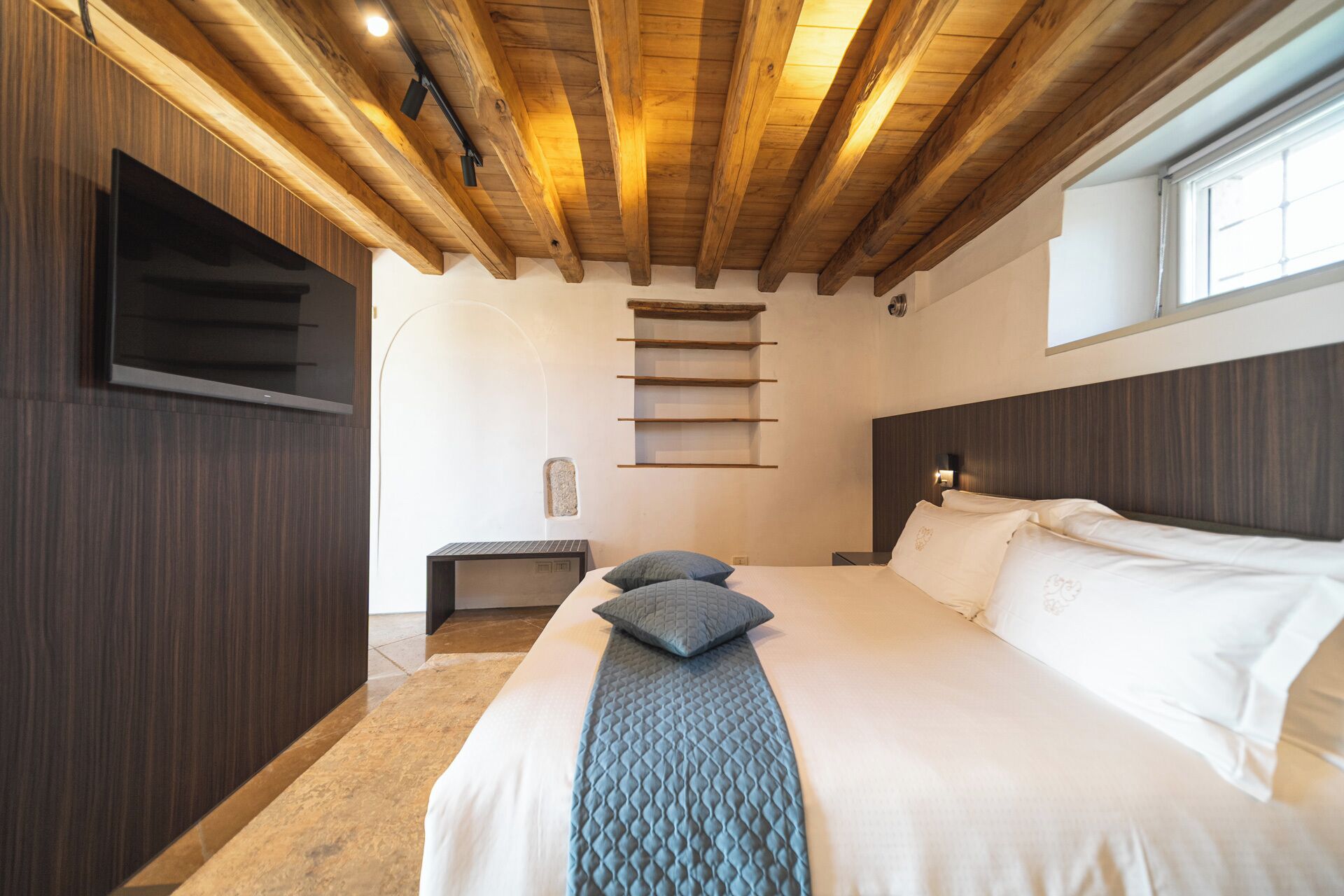 Un dormitorio con techo de madera, una cama con ropa blanca y almohadas azules, un televisor montado y una ventana con luz natural.