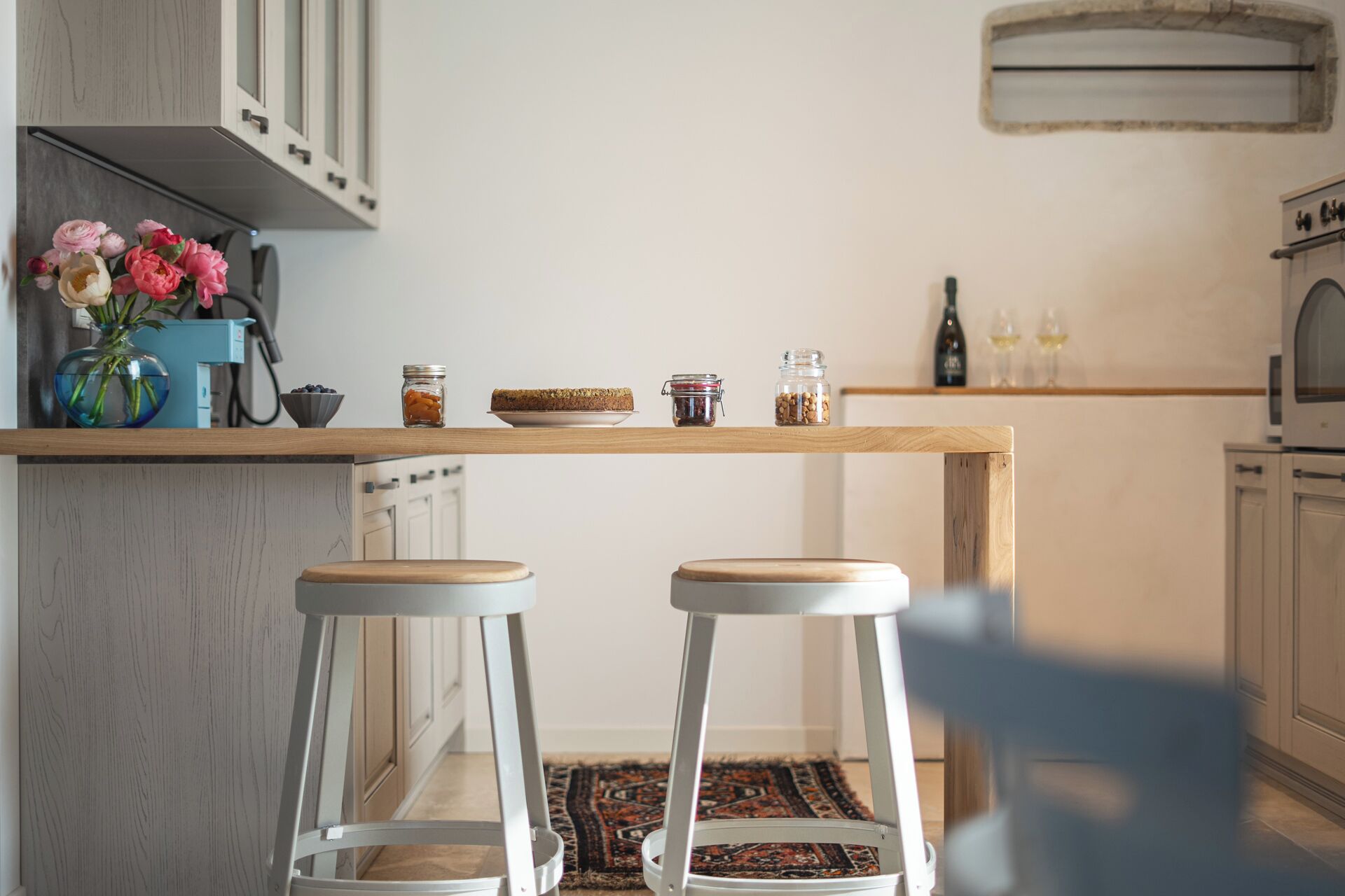 A kitchen with a wooden countertop, two white stools, a vase with flowers, and jars of food.