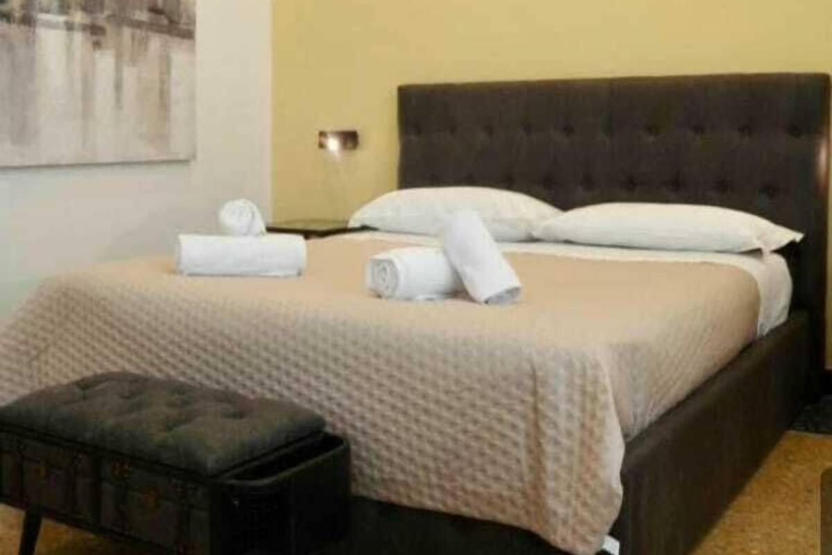 Una habitación de hotel con una cama grande, cabecera oscura y un pequeño reposapiés negro.