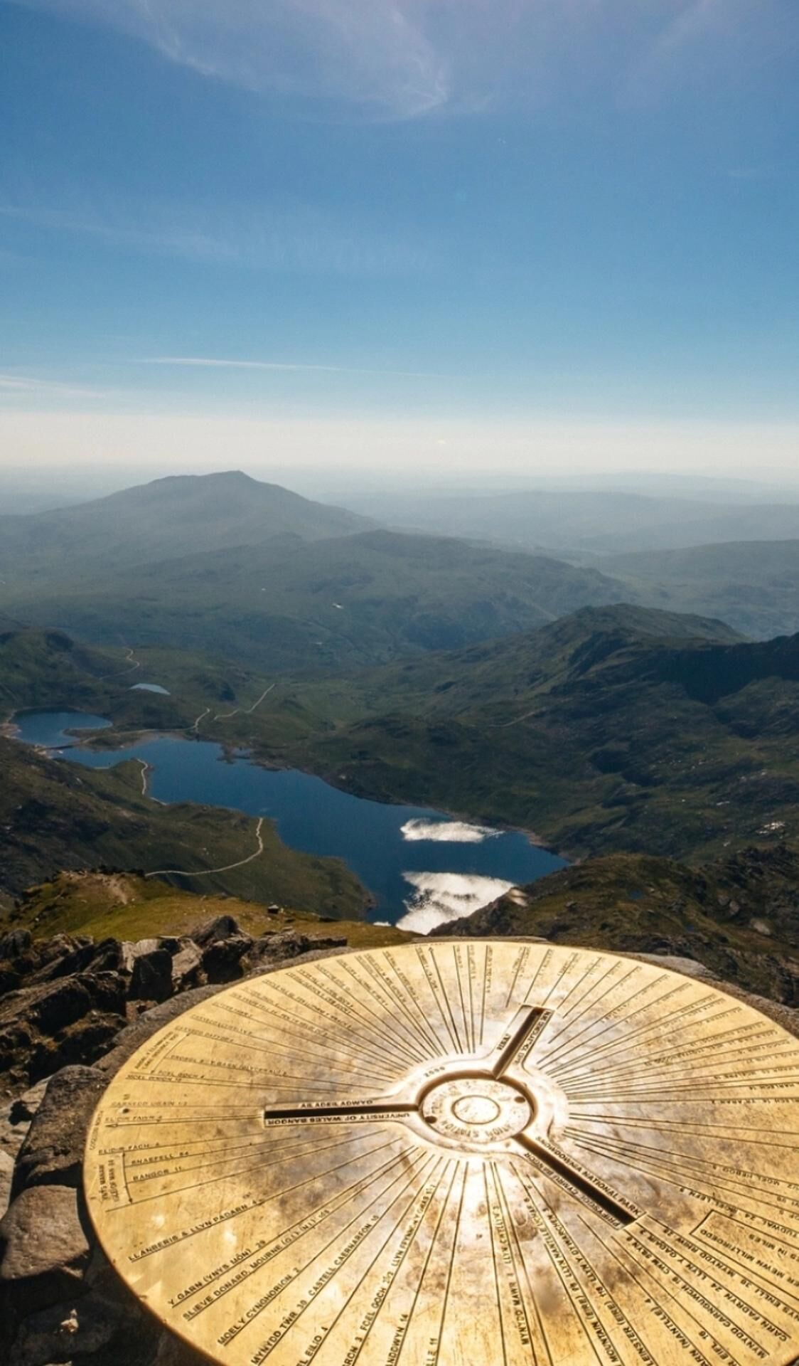 Un monument circulaire en pierre, orné d’inscriptions, entouré d’un paysage montagneux avec un lac et un ciel dégagé.
