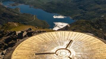 Un monument circulaire en pierre, orné d’inscriptions, entouré d’un paysage montagneux avec un lac et un ciel dégagé.