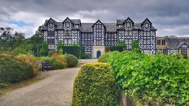 En traditionell byggnad i tudorstil med en symmetrisk fasad, en central entré och en grusgång som leder fram till en bänk.