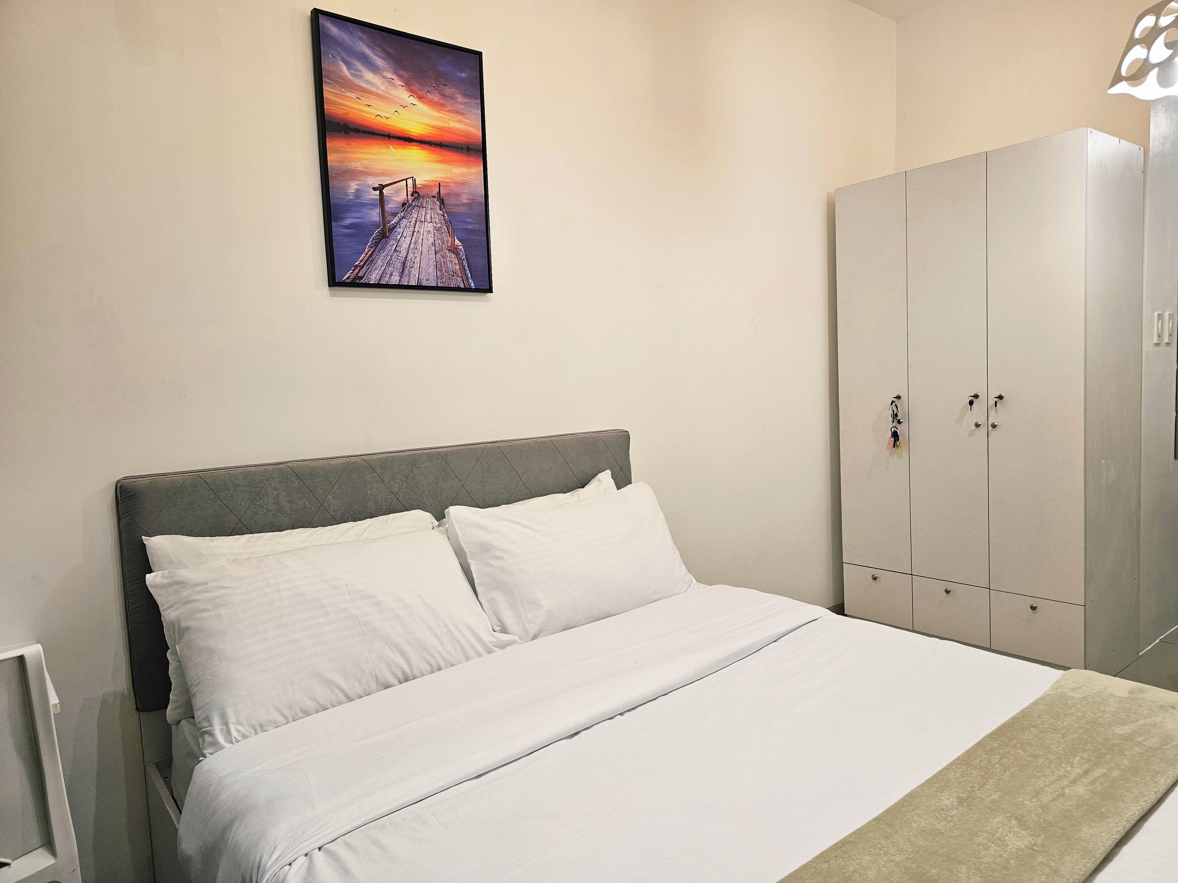A bedroom with a bed, white bedding, a grey headboard, a wardrobe, and a sunset painting on the wall.