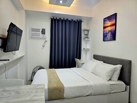 Habitación de hotel con cama, escritorio con computadora, televisión de pantalla plana, aire acondicionado, ventana con cortinas oscuras y un cuadro en la pared.