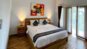 A bedroom with a wooden bed, white bedding, a nightstand with a lamp, a wall-mounted painting, and a window with curtains. - Green Sunrise Hill View (Penida Island)