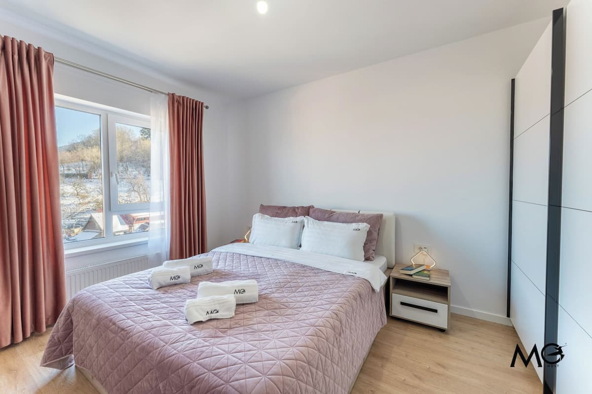 A modern bedroom with a large bed, a bedside table with a lamp, and a view of the outside.