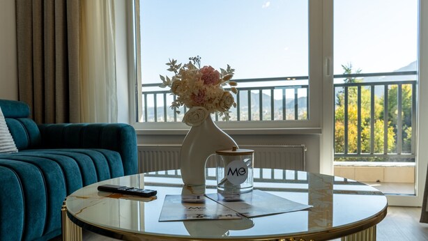 Una habitación con un sofá azul turquesa, una mesa de centro redonda de vidrio con un control remoto, un florero con flores y vistas a un balcón con barandilla y árboles.