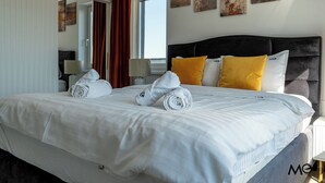 Una cama bien tendida con sábanas blancas y toallas enrolladas, un cabecero oscuro y dos almohadas amarillas.