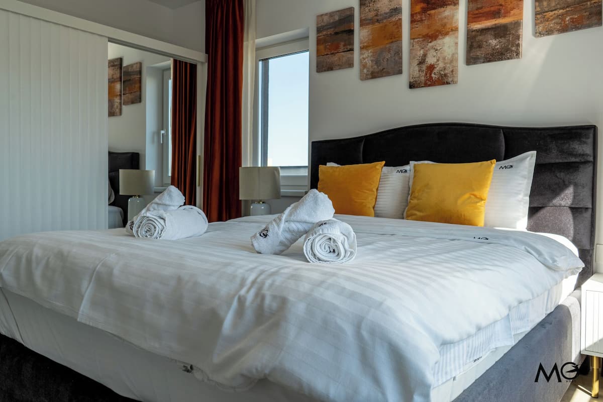 A neatly made bed with white linens and rolled towels, a dark headboard, and two yellow pillows.