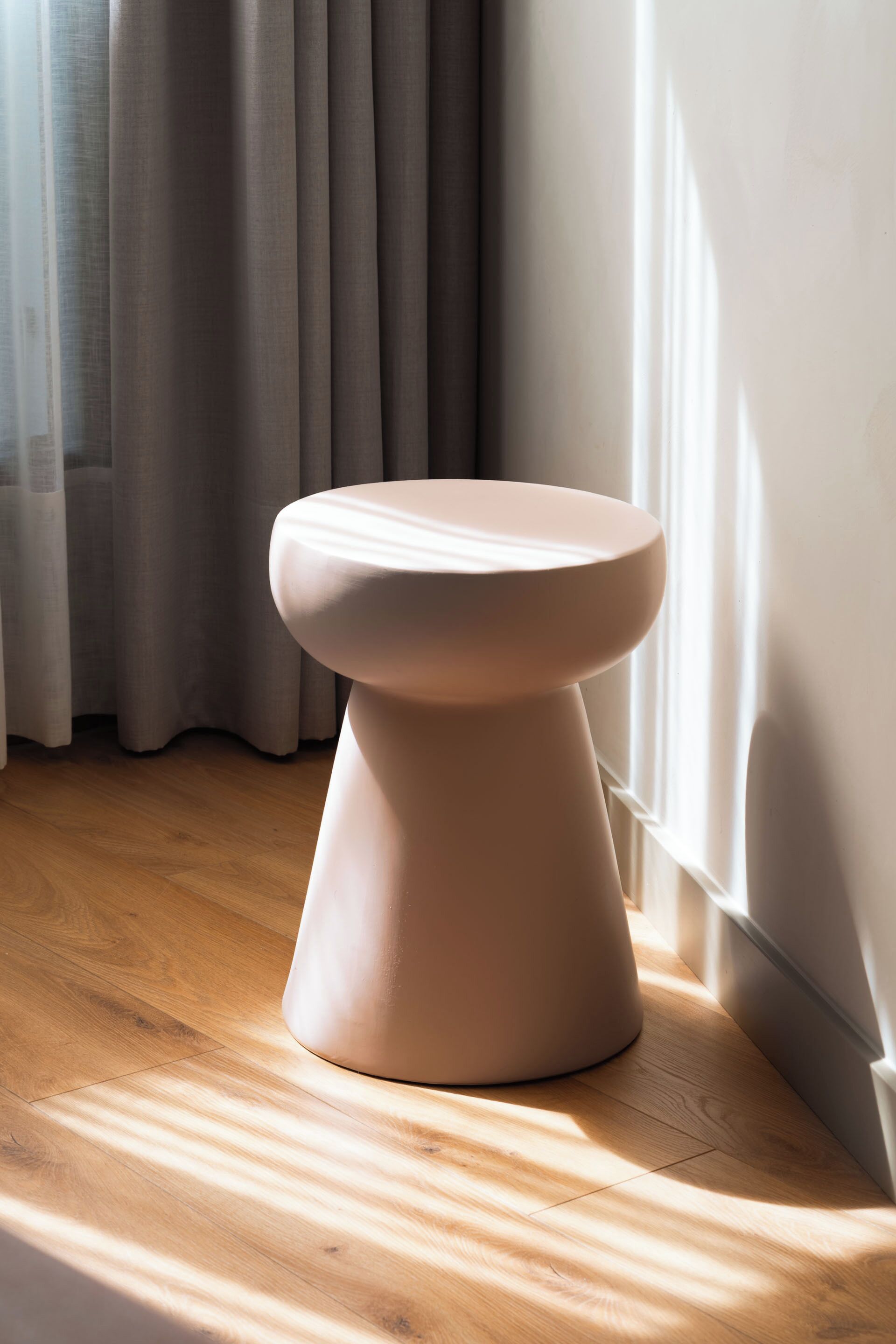 A modern stool in a corner of a room with wooden flooring and a window with curtains.