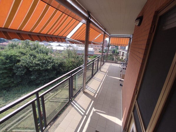 A balcony with a view of greenery and buildings, a tiled floor, and a metal railing.