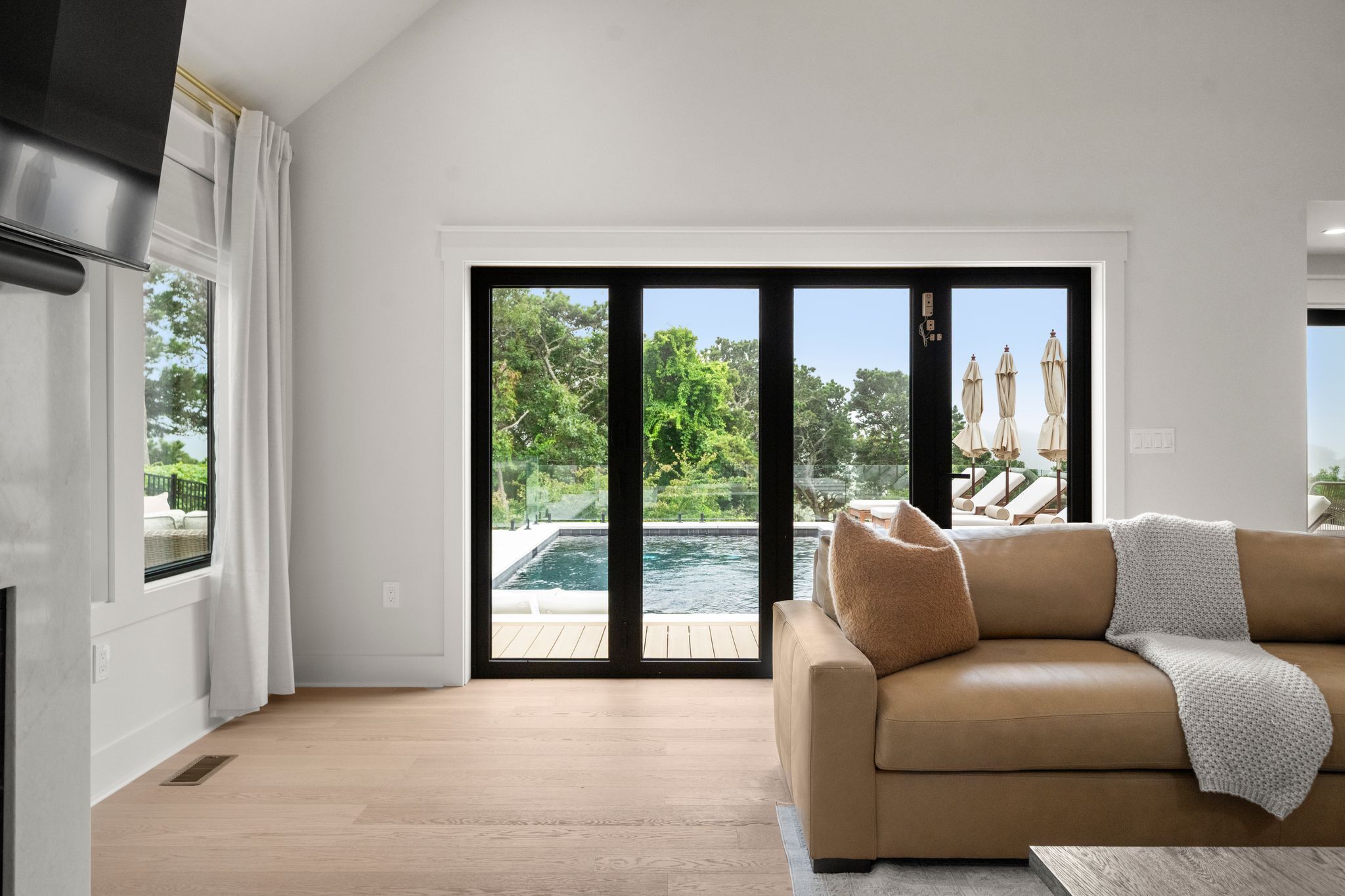 A modern living room with a beige sofa, a large sliding glass door leading to a pool, and a view of lush greenery.
