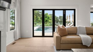 A modern living room with a beige sofa, a large sliding glass door leading to a pool, and a view of lush greenery.