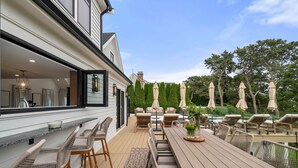 An outdoor patio with a wooden table and wicker chairs, a pool area with lounge chairs, and a view of the surrounding greenery.