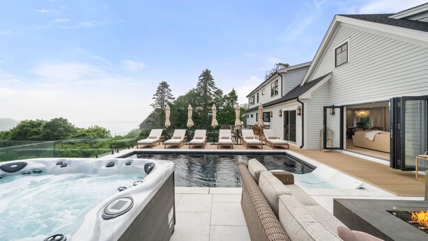 A modern outdoor pool area with a hot tub, lounge chairs, and a deck overlooking a residential property.