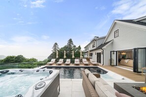 Un área de piscina moderna al aire libre con jacuzzi, sillones de descanso y una terraza con vistas a una propiedad residencial.