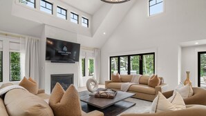 A spacious living room with a high ceiling, a fireplace, a large flat-screen TV, and a comfortable seating area with sofas and cushions.