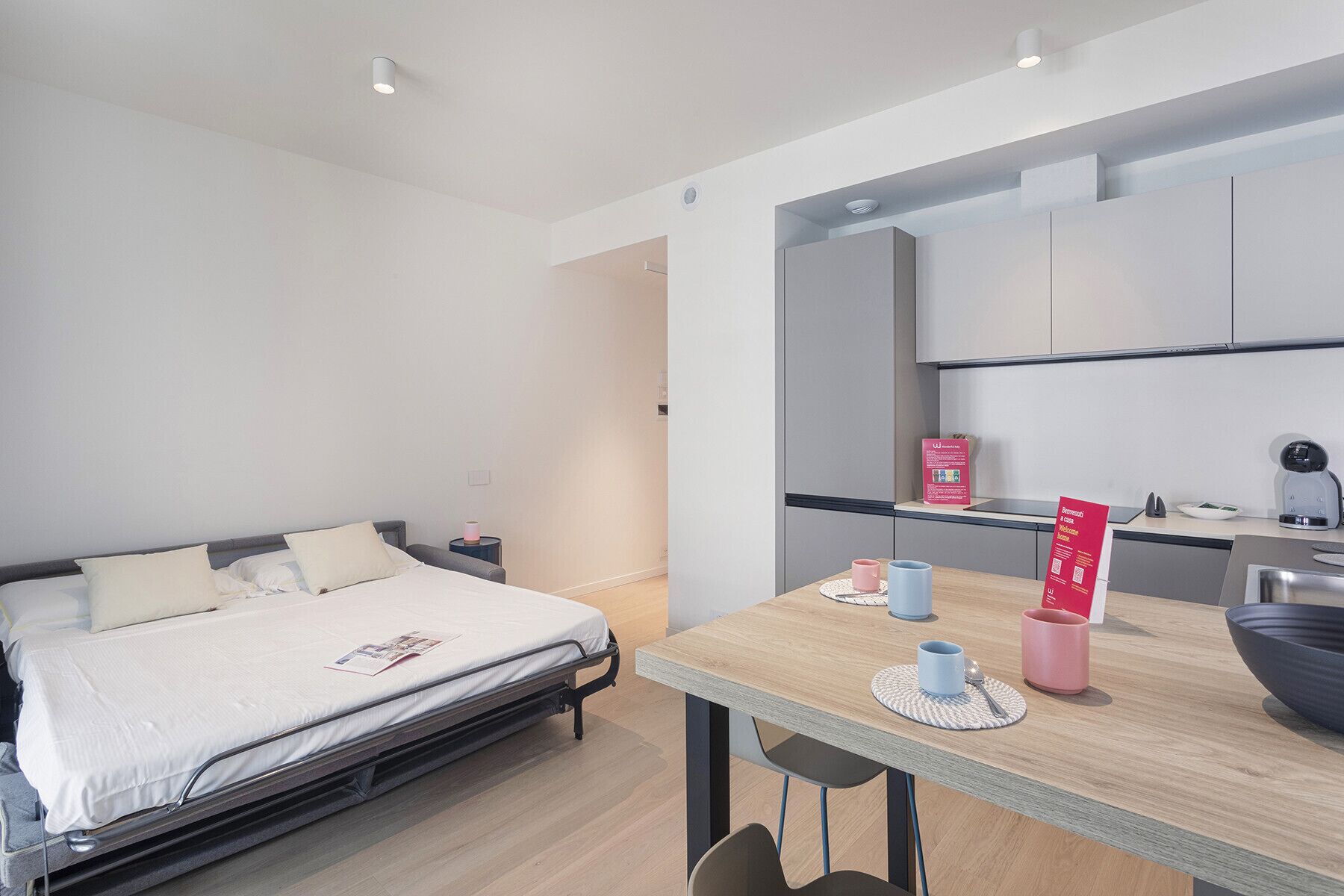 Et moderne hotelværelse med seng, et lille bord med kopper og et skilt, samt et tekøkken i baggrunden.