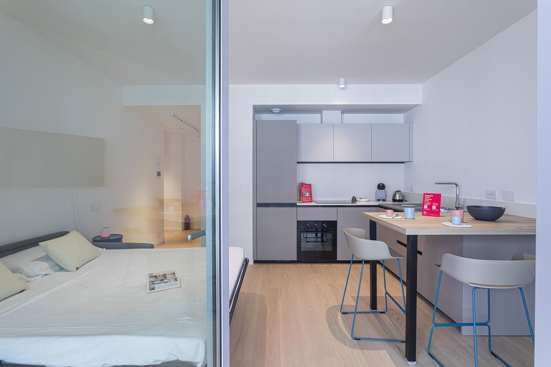 Una habitación de hotel moderna con cama, kitchenette pequeña y zona de comedor con taburetes.