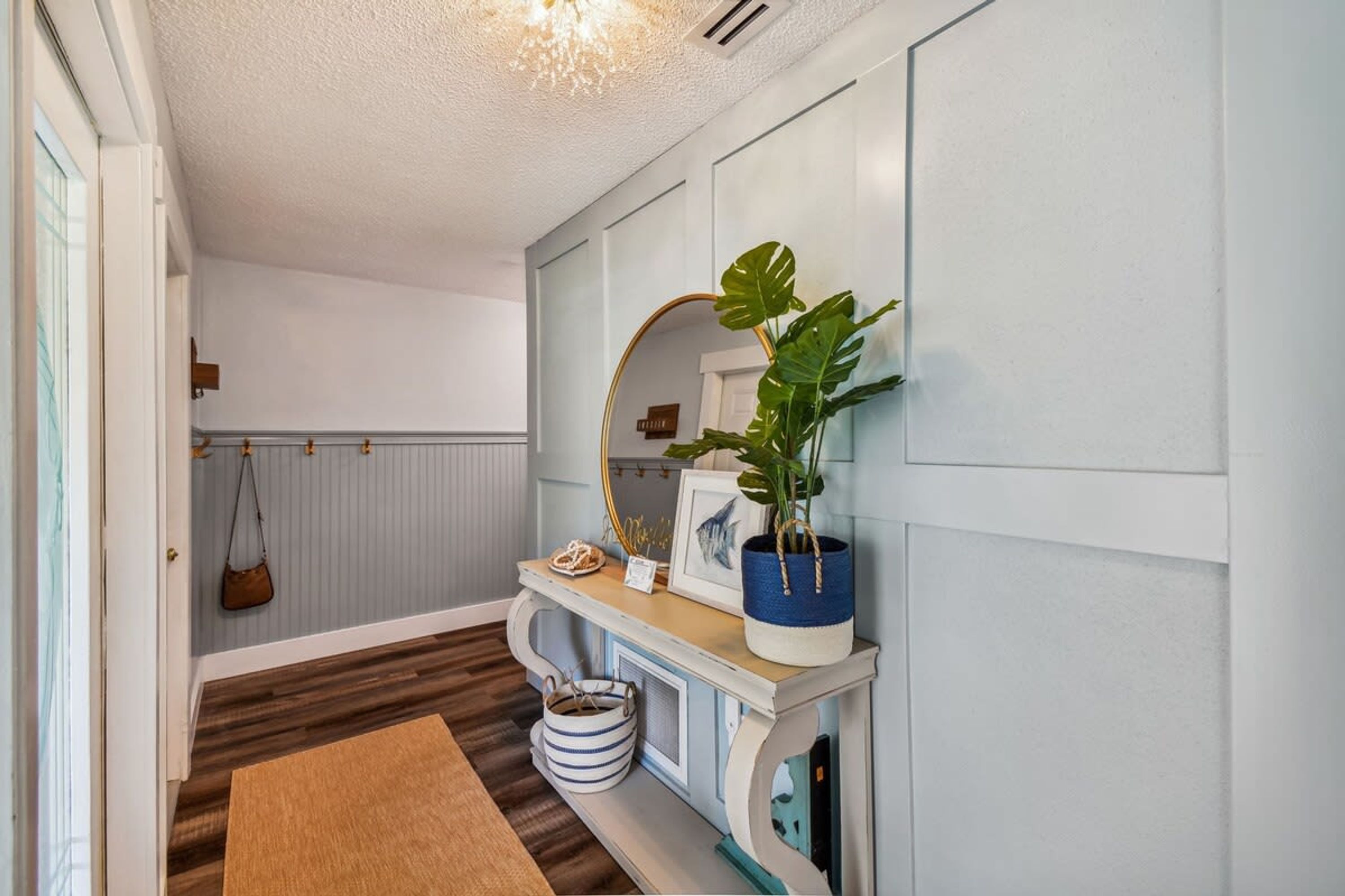 A hallway with a wooden floor, a white door, a mirror, a plant in a blue pot, and a small table with a framed picture and a handbag hanging on the wall.