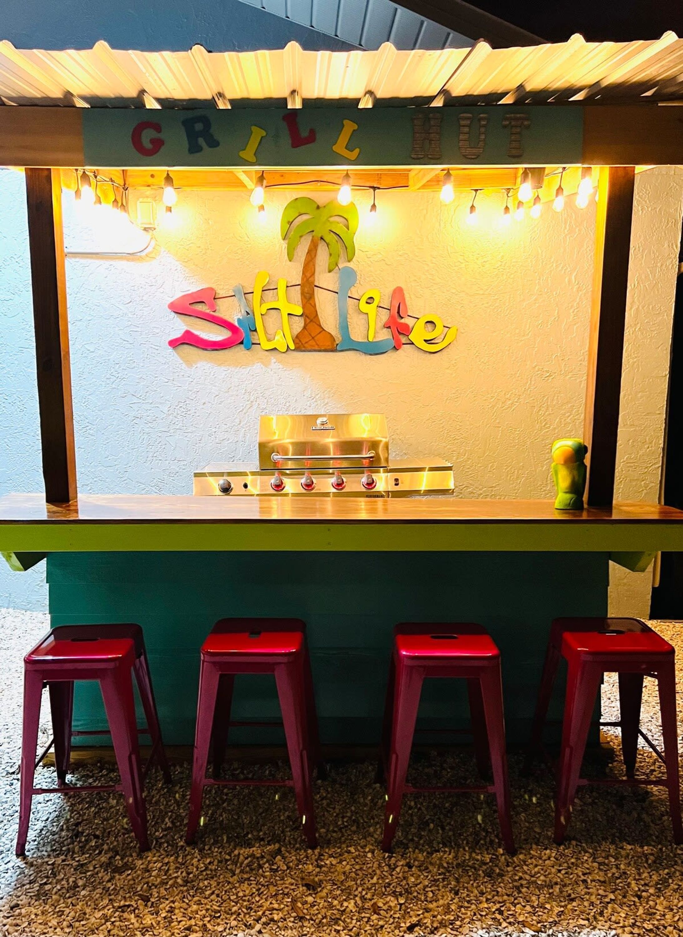 A small outdoor grill with a colorful sign reading "Sitt Lite" and a palm tree decoration. There are four red stools in front.